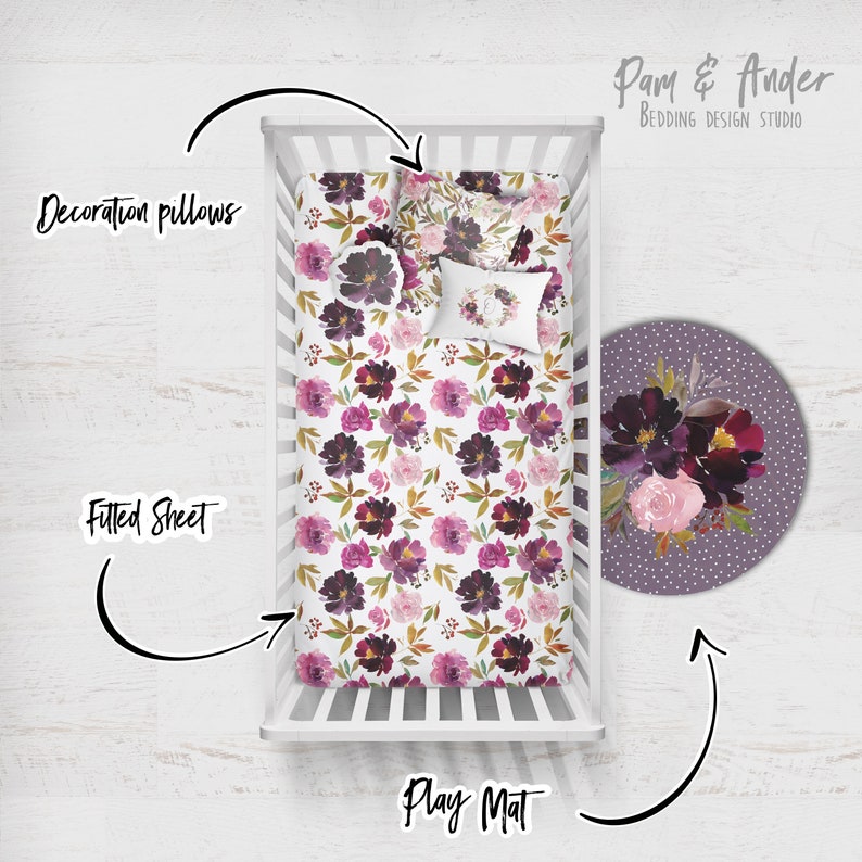 May include: A white cot with a floral patterned fitted sheet and two decorative pillows. A round play mat with a floral design is on the floor next to the cot. The text "Pam & Ander Bedding Design Studio" is in the top right corner of the image.