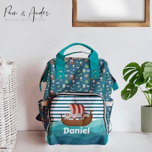 Może przedstawiać: Torba na pieluchy w kolorze turkusowym i zielonym z kieszenią z przodu, na której znajduje się kreskówkowa ilustracja statku wikingów z etykietą z imieniem "Daniel".