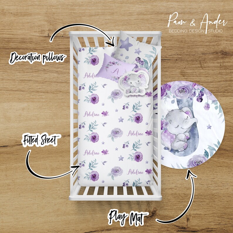 May include: A white cot with a purple and white floral fitted sheet and a matching play mat. The play mat features a watercolour illustration of a sleeping elephant with purple flowers. The cot also includes a decorative pillow with the name "Adeline" on it.