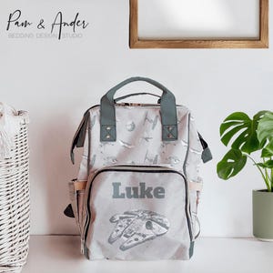 May include: A gray and white diaper bag with a Star Wars theme. The bag has a large image of the Millennium Falcon and the name "Luke" printed on the front.