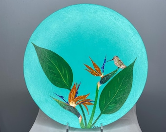 Può includere: Un piatto rotondo in legno dipinto di color turchese con un uccello e fiori di uccello del paradiso dipinti su di esso. L'uccello è appollaiato su una foglia e i fiori sono in fiore. Il piatto è su un supporto bianco.