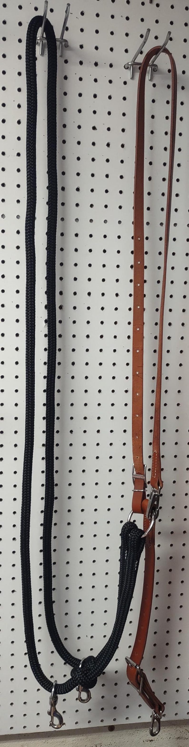 May include: A black rope halter and a brown leather horse rein hang against a white pegboard. The halter has a knot and metal hardware. The leather rein has a buckle and metal hardware.