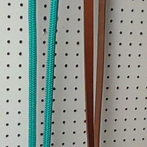May include: A turquoise rope and a brown leather strap hang against a white pegboard. The rope is looped at the bottom with a metal clasp. The leather strap has a metal clasp and a loop.