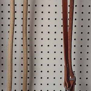 May include: A tan rope and brown leather horse rein set. The rope is braided and attached to a leather strap with metal hardware. The set is designed for equestrian use.