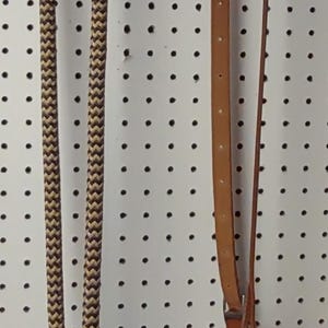 May include: A brown and tan rope halter and leather reins hang against a white pegboard background. The rope has a chevron pattern. The leather reins are a medium brown color and have metal hardware.