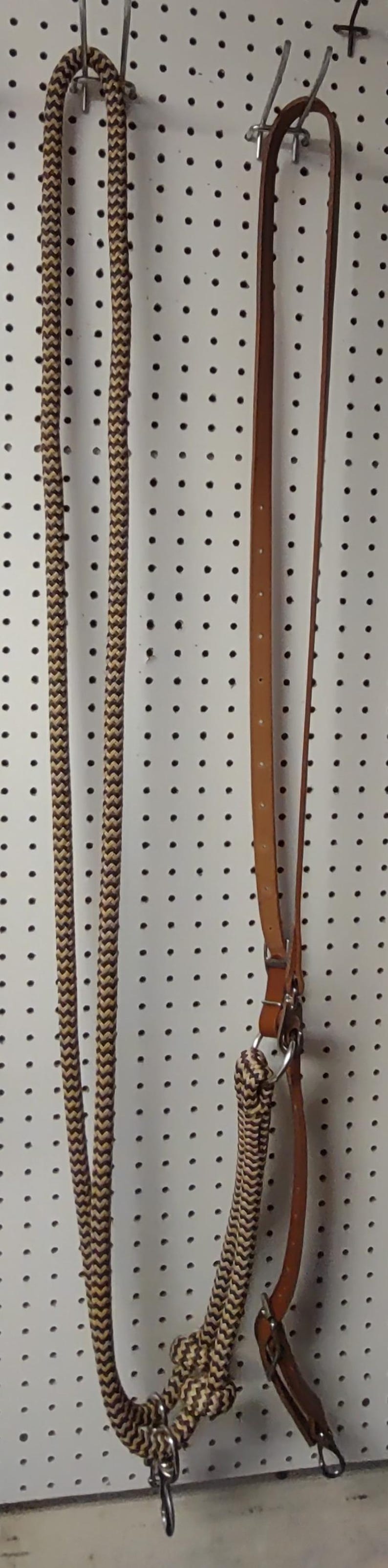 May include: A brown and tan rope halter and leather reins hang against a white pegboard background. The rope has a chevron pattern. The leather reins are a medium brown color and have metal hardware.