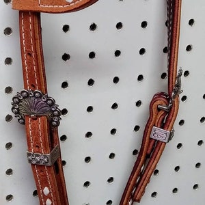 May include: A brown leather horse headstall with white stitching and silver conchos. The headstall is hanging on a pegboard with holes.