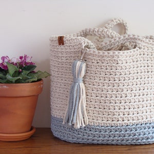 Puede incluir: Un bolso de mano de crochet de color azul claro y blanco. El bolso tiene un borla y una etiqueta de cuero. El bolso est&aacute; sobre una superficie de madera junto a una planta en maceta.
