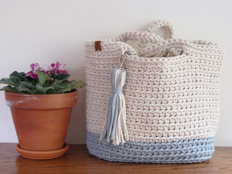 Puede incluir: Un bolso de mano de crochet de color azul claro y blanco. El bolso tiene un borla y una etiqueta de cuero. El bolso est&aacute; sobre una superficie de madera junto a una planta en maceta.