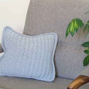 May include: A light blue knitted throw pillow with a herringbone pattern sits on a gray armchair with a wooden armrest. A green plant is visible in the background.