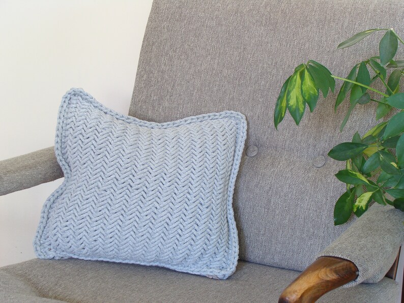May include: A light blue knitted throw pillow with a herringbone pattern sits on a gray armchair with a wooden armrest. A green plant is visible in the background.