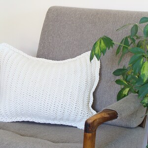 May include: A white crocheted pillow with a herringbone pattern sits on a gray armchair with a wooden armrest. A green leafy plant is in the background.
