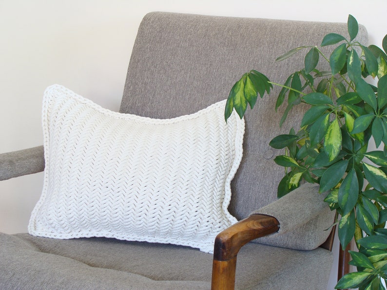 May include: A white crocheted pillow with a herringbone pattern sits on a gray armchair with a wooden armrest. A green leafy plant is in the background.