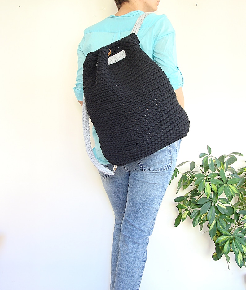 May include: A black crocheted backpack with a drawstring closure and gray straps. The backpack is being worn by a person wearing a light blue shirt and blue jeans.