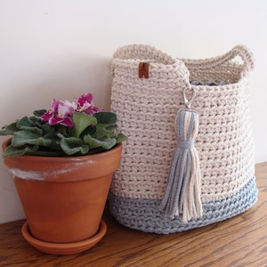 Puede incluir: Una cesta de almacenamiento de crochet en un esquema de color crema y azul claro. La cesta tiene un borla y est&aacute; sentada sobre una superficie de madera junto a una planta en maceta con flores moradas.