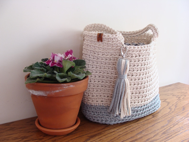 Puede incluir: Una cesta de almacenamiento de crochet en un esquema de color crema y azul claro. La cesta tiene un borla y est&aacute; sentada sobre una superficie de madera junto a una planta en maceta con flores moradas.