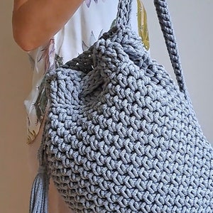 May include: A gray crocheted backpack with a drawstring closure and a tassel. The backpack is made of thick rope and has a simple, minimalist design.
