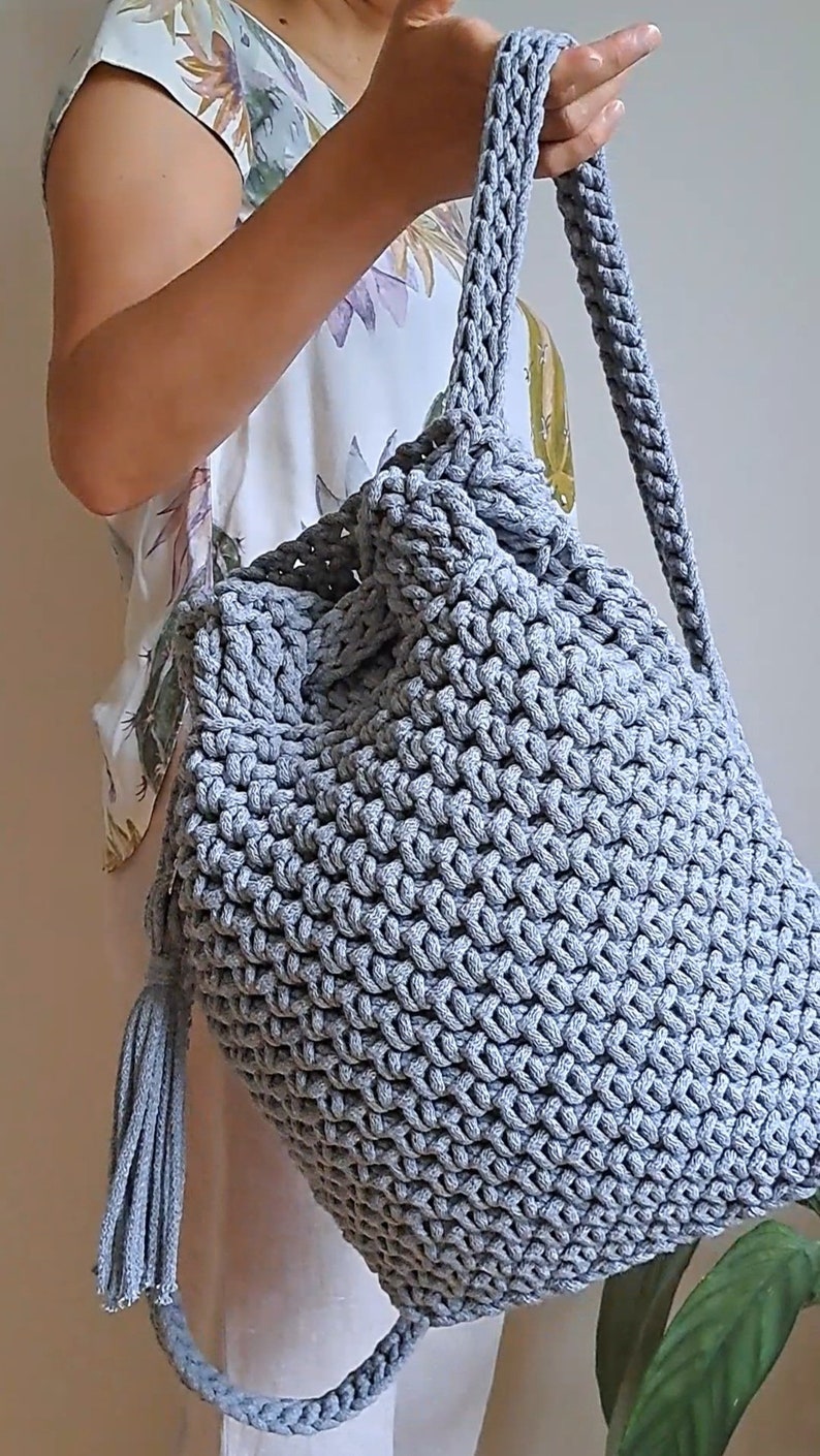 May include: A gray crocheted backpack with a drawstring closure and a tassel. The backpack is made of thick rope and has a simple, minimalist design.