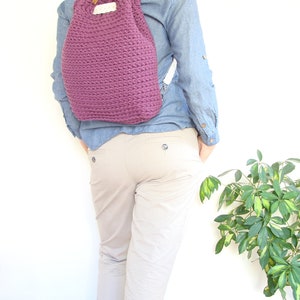 May include: A purple crocheted drawstring backpack with white straps. The backpack is being worn by a person wearing a blue denim shirt and beige pants.
