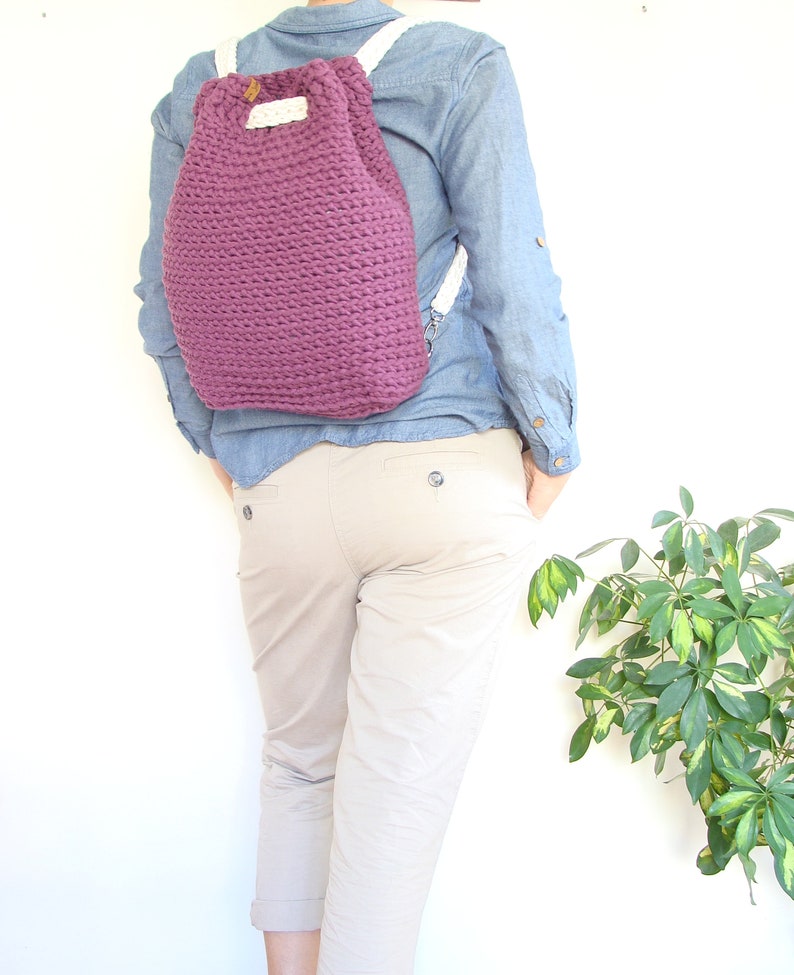 May include: A purple crocheted drawstring backpack with white straps. The backpack is being worn by a person wearing a blue denim shirt and beige pants.