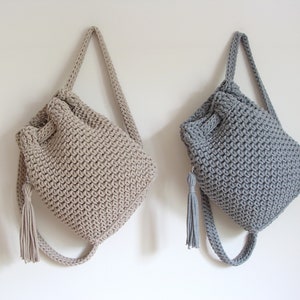 May include: Two crocheted drawstring bags with long straps and tassels. One bag is beige and the other is gray.