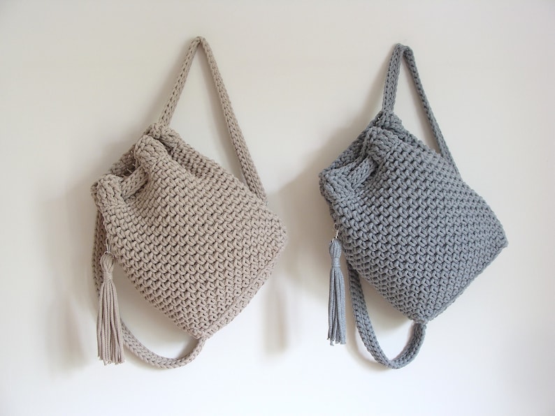 May include: Two crocheted drawstring bags with long straps and tassels. One bag is beige and the other is gray.