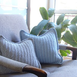 May include: Two light blue throw pillows with a herringbone pattern are placed on a gray armchair. The pillows are made of a soft, textured fabric and have a subtle sheen.