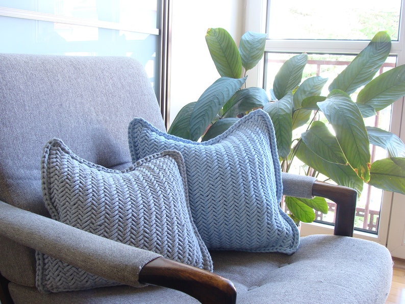 May include: Two light blue throw pillows with a herringbone pattern are placed on a gray armchair. The pillows are made of a soft, textured fabric and have a subtle sheen.
