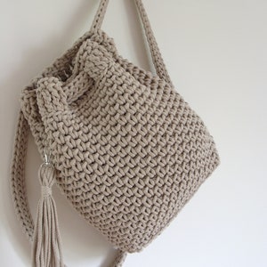 May include: A beige crocheted backpack with a long tassel hanging from the bottom. The backpack has a drawstring closure and adjustable straps.