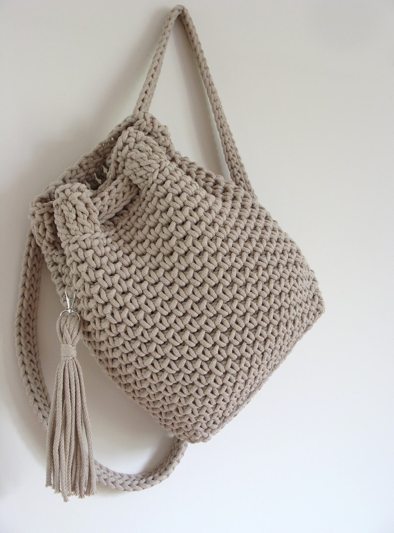 May include: A beige crocheted backpack with a long tassel hanging from the bottom. The backpack has a drawstring closure and adjustable straps.