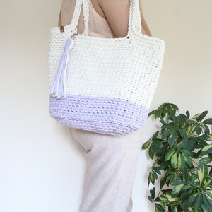 Puede incluir: Una bolsa de mano de crochet blanca y lavanda con un borla blanca. La bolsa tiene dos asas y est&aacute; hecha de hilo grueso.