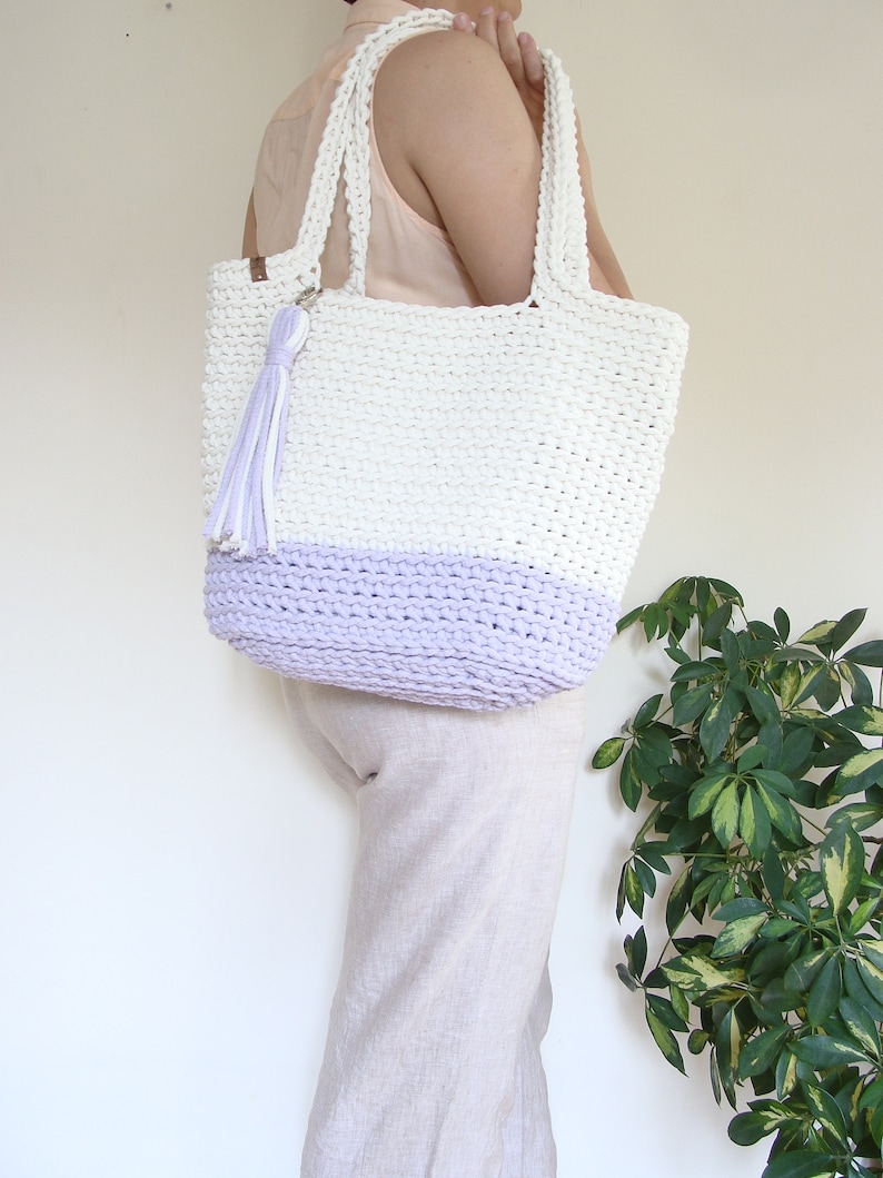 Puede incluir: Una bolsa de mano de crochet blanca y lavanda con un borla blanca. La bolsa tiene dos asas y est&aacute; hecha de hilo grueso.