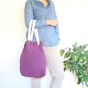 May include: A purple crocheted tote bag with white braided handles. The bag is being held by a person wearing a light blue denim shirt and beige pants.