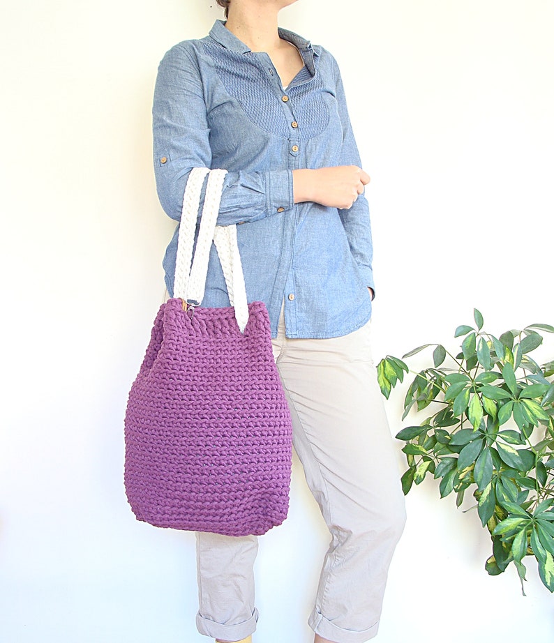 May include: A purple crocheted tote bag with white braided handles. The bag is being held by a person wearing a light blue denim shirt and beige pants.