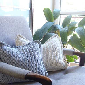May include: Two knitted throw pillows in a herringbone pattern, one in light gray and one in white, are placed on a gray armchair. The armchair is in front of a window with a green plant in the background.
