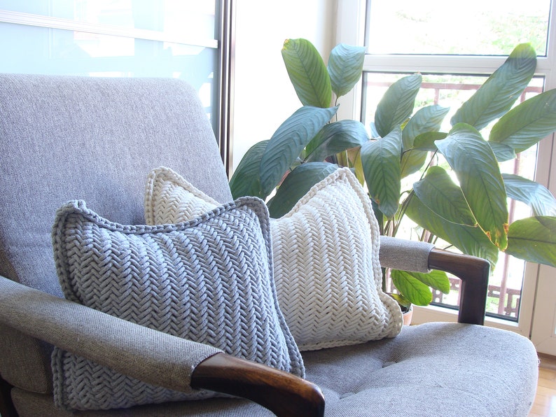 May include: Two knitted throw pillows in a herringbone pattern, one in light gray and one in white, are placed on a gray armchair. The armchair is in front of a window with a green plant in the background.