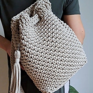 May include: A beige crocheted backpack with a long tassel. The backpack is being worn by a person.