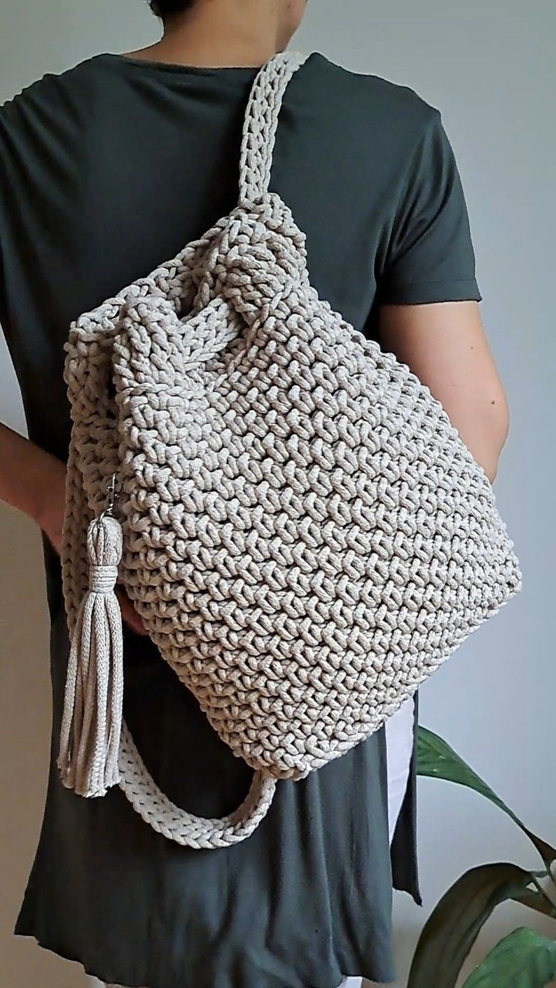 May include: A beige crocheted backpack with a long tassel. The backpack is being worn by a person.