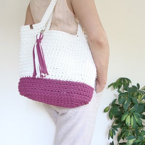 Puede incluir: Bolso tote de crochet blanco con fondo de crochet rosa y un borla blanco y rosa. El bolso lo lleva una persona.