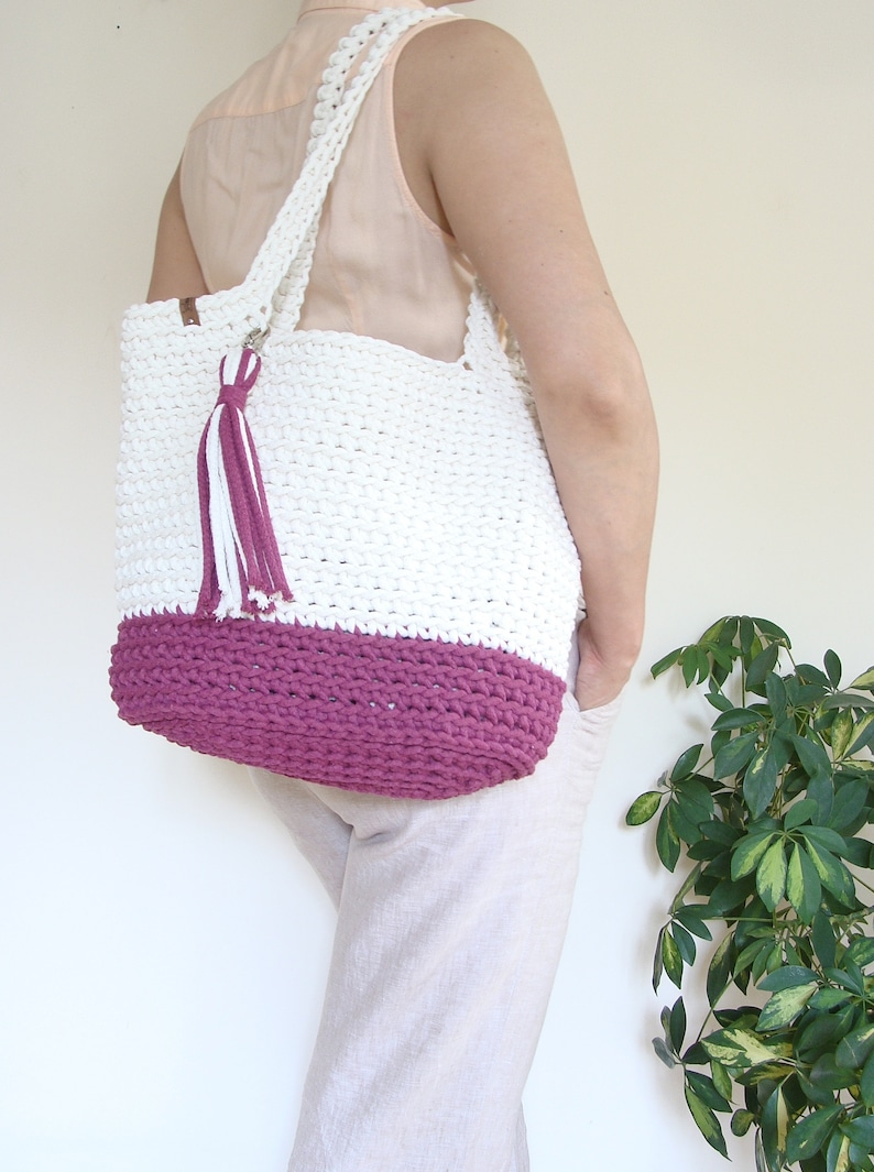 Puede incluir: Bolso tote de crochet blanco con fondo de crochet rosa y un borla blanco y rosa. El bolso lo lleva una persona.