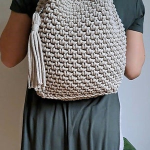May include: A beige crocheted backpack with a drawstring closure and a tassel. The backpack is being worn by a person.