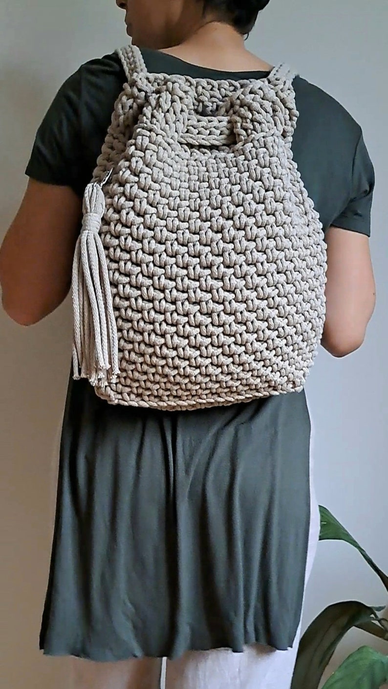 May include: A beige crocheted backpack with a drawstring closure and a tassel. The backpack is being worn by a person.