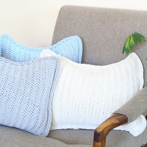 May include: Three knitted throw pillows in a herringbone pattern. The pillows are light blue, white, and a darker shade of blue. The pillows are arranged on a gray armchair with a wooden armrest.