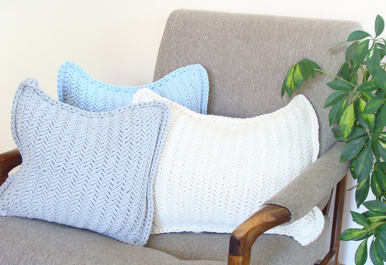 May include: Three knitted throw pillows in a herringbone pattern. The pillows are light blue, white, and a darker shade of blue. The pillows are arranged on a gray armchair with a wooden armrest.