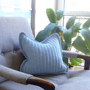 May include: A light blue knitted pillow with a herringbone pattern sits on a gray armchair with a wooden armrest. The armchair is in front of a window with a green plant.