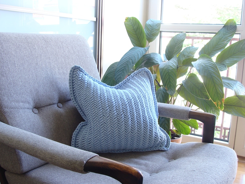 May include: A light blue knitted pillow with a herringbone pattern sits on a gray armchair with a wooden armrest. The armchair is in front of a window with a green plant.