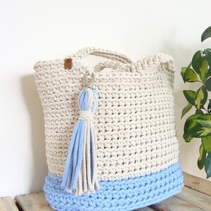 Puede incluir: Un bolso de mano de crochet en color beige y azul claro. El bolso tiene un borla azul claro atada al asa.