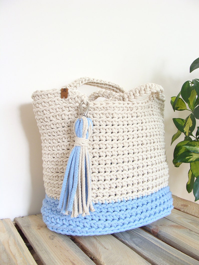 Puede incluir: Un bolso de mano de crochet en color beige y azul claro. El bolso tiene un borla azul claro atada al asa.