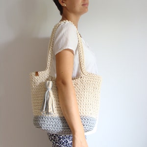 Puede incluir: Un bolso de mano de crochet de color crema y gris. El bolso tiene una correa larga y un borla.