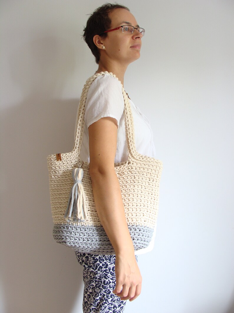 Puede incluir: Un bolso de mano de crochet de color crema y gris. El bolso tiene una correa larga y un borla.